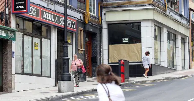 Dos vecinos pasean por la calle de La Argañosa con bolsas en la mano con la estampa que adorna el barrio de Oviedo, negocios cerrados con carteles buscando dueño. / ALEX PIÑA